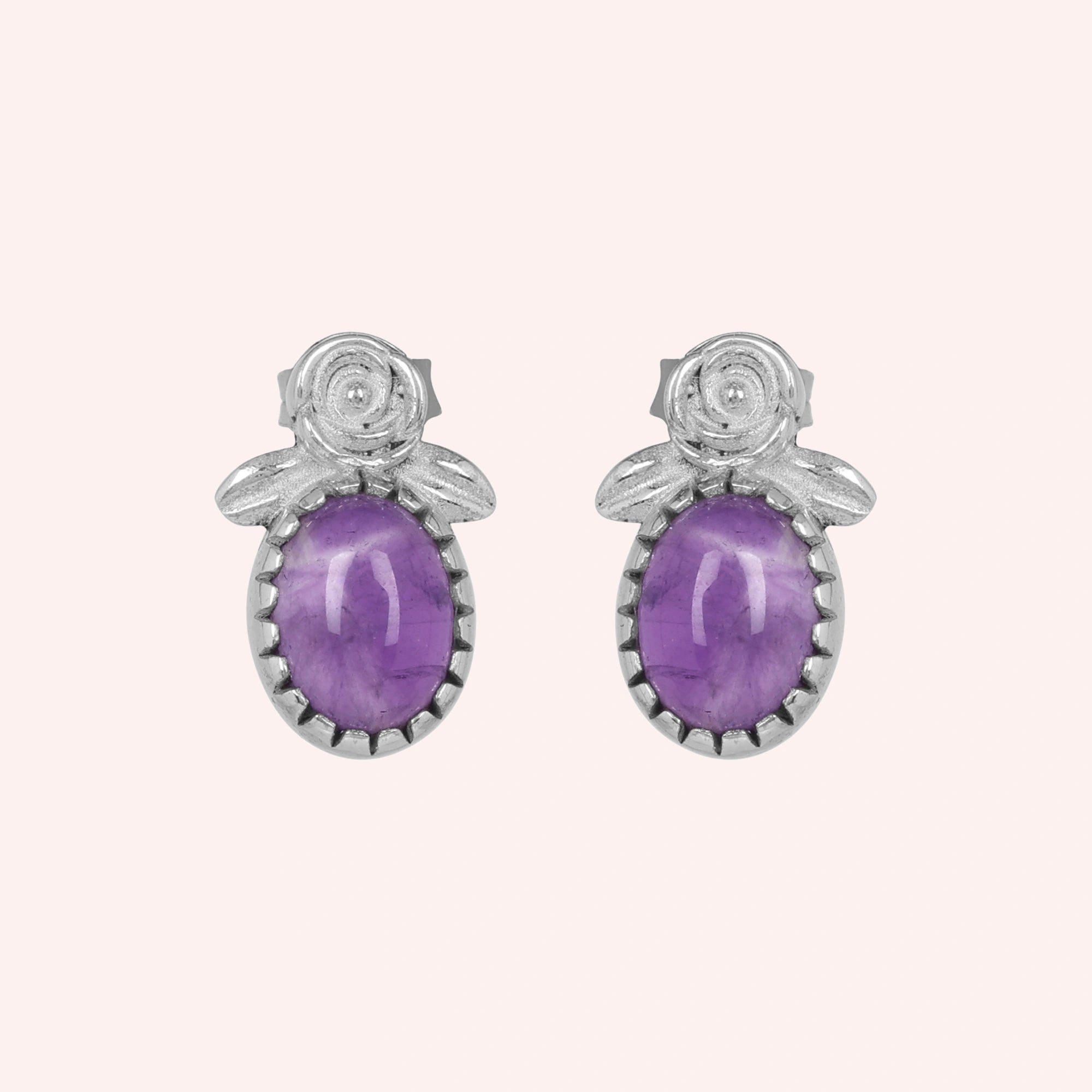 Front_view_amethyst_stud_earrings_with_oval_purple_gemstone_set_in_silver_flower_stud_earrings_displayed_on_plain_pink_background