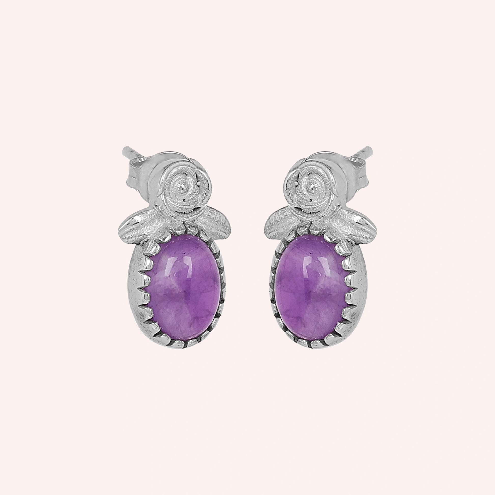 Front_view_amethyst_stud_earrings_with_oval_purple_gemstone_set_in_silver_flower_stud_earrings_shown_on_soft_pink_background