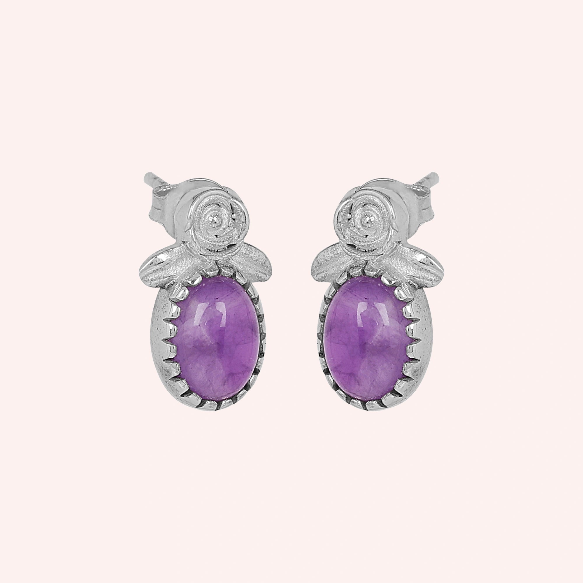 Front_view_amethyst_stud_earrings_with_oval_purple_gemstone_set_in_silver_flower_stud_earrings_shown_on_soft_pink_background
