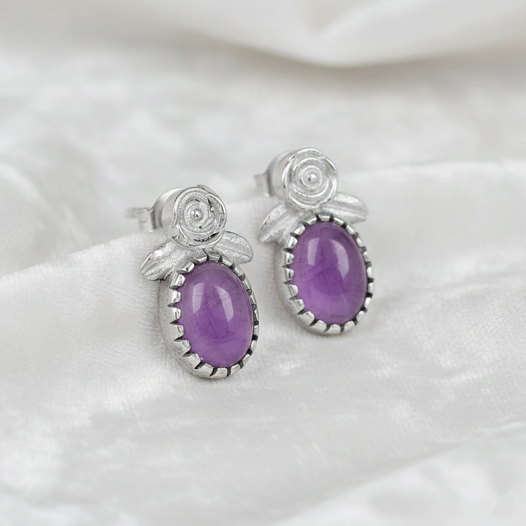 Angled_front_view_amethyst_stud_earrings_with_oval_purple_gemstone_set_in_silver_flower_stud_earrings_on_white_fabric_background