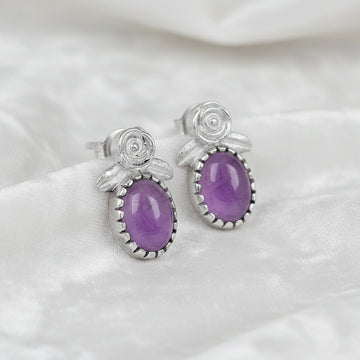 Angled_front_view_amethyst_stud_earrings_with_oval_purple_gemstone_set_in_silver_flower_stud_earrings_on_white_fabric_background