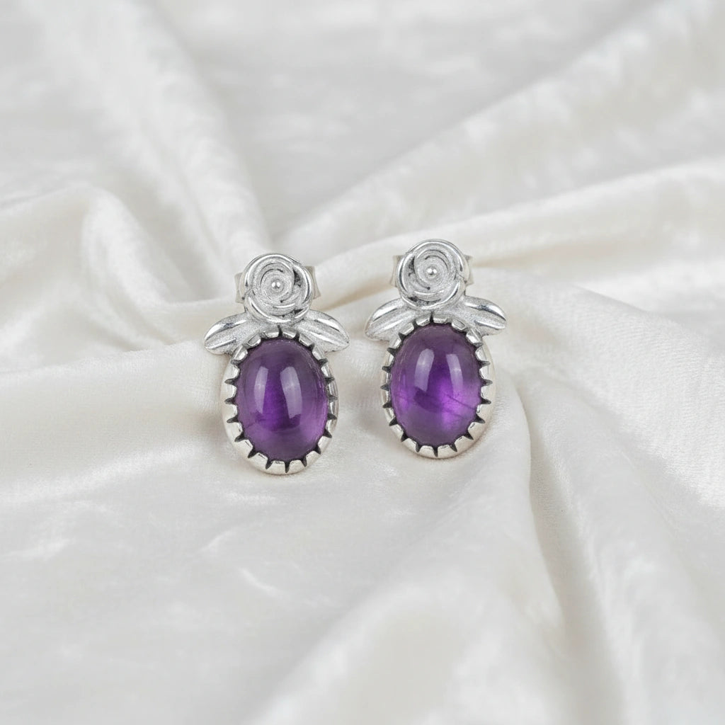 Front_view_amethyst_stud_earrings_with_oval_purple_gemstone_set_in_silver_flower_stud_earrings_on_white_fabric_background
