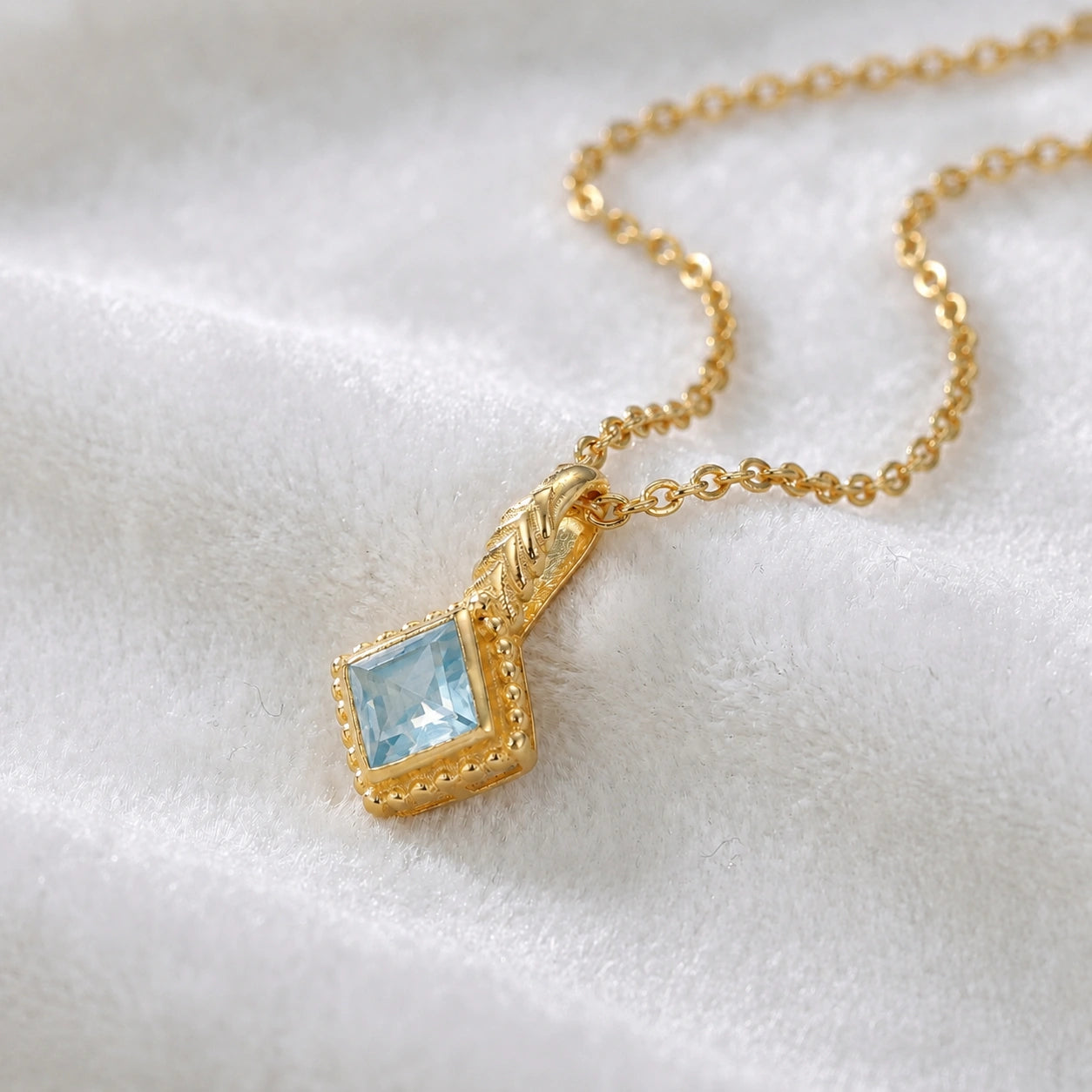 Birthstone_necklace_in_gold_plated_silver_with_square_cut_aquamarine_pendant_in_slighlty_angled_view_on_soft_white_fabric_background