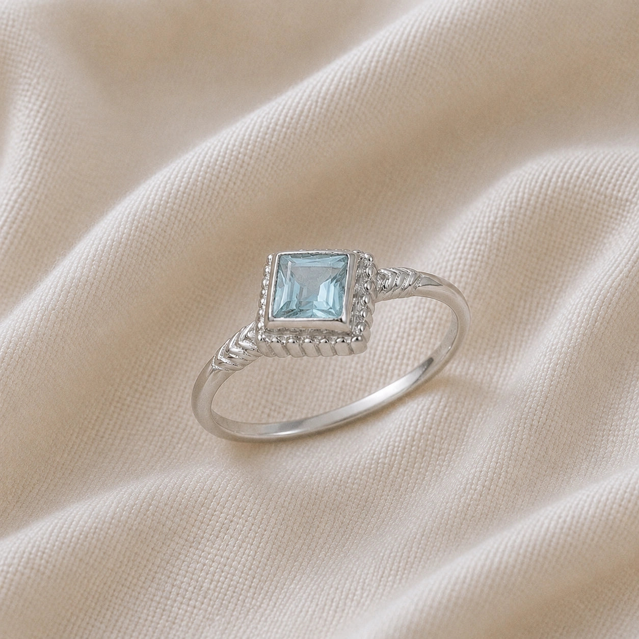 Princess_cut_ring_sterling_silver_with_square_aquamarine_and_detailed_band_slightly_tilted_view_on_soft_fabric_background