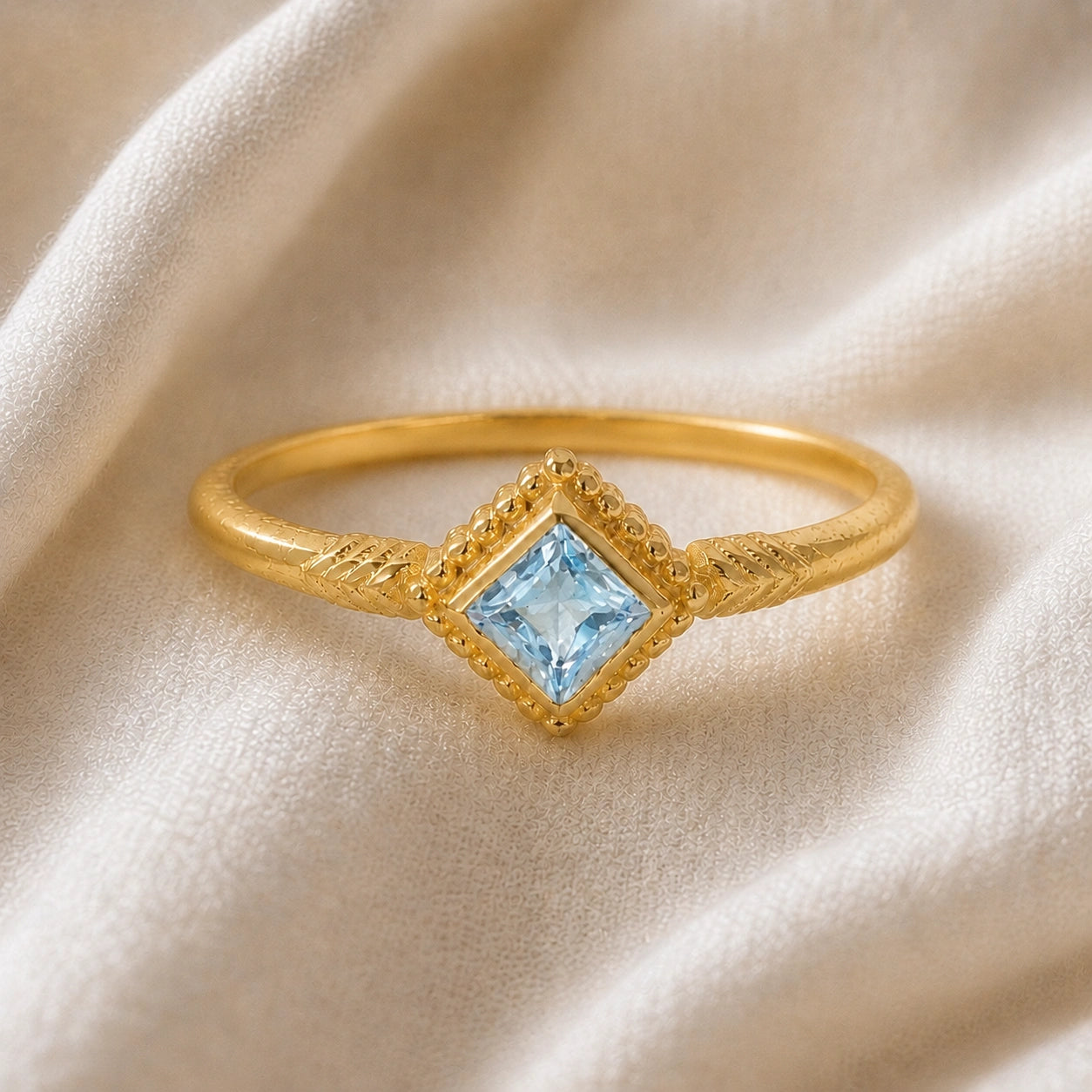Princess_cut_ring_gold_plated_silver_with_square_aquamarine_and_detailed_band_front_angle_view_on_soft_fabric_background