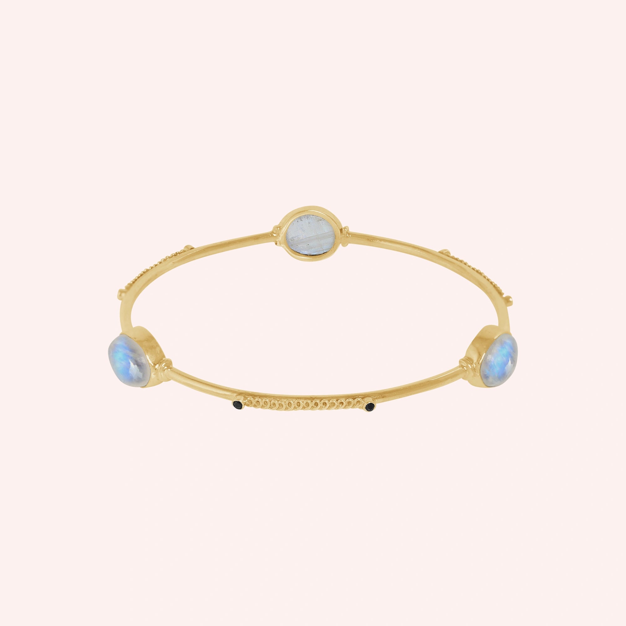 Bangle_bracelet_in_gold_plated_silver_with_oval_moonstones_and_black_onyx_stones_at_front_angle_view_on_solid_pink_background