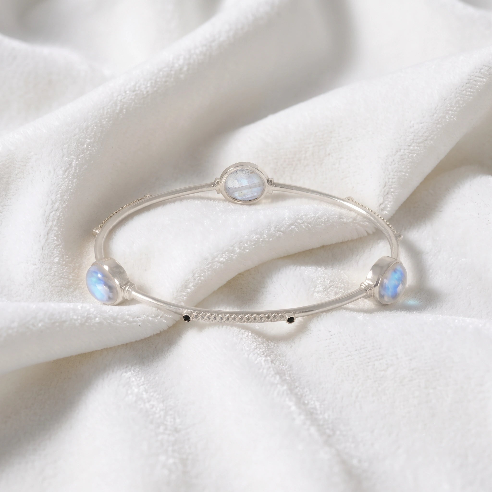 Bangle_bracelet_in_sterling_silver_with_oval_moonstones_and_black_onyx_stones_at_front_angle_view_on_soft_fabric_background