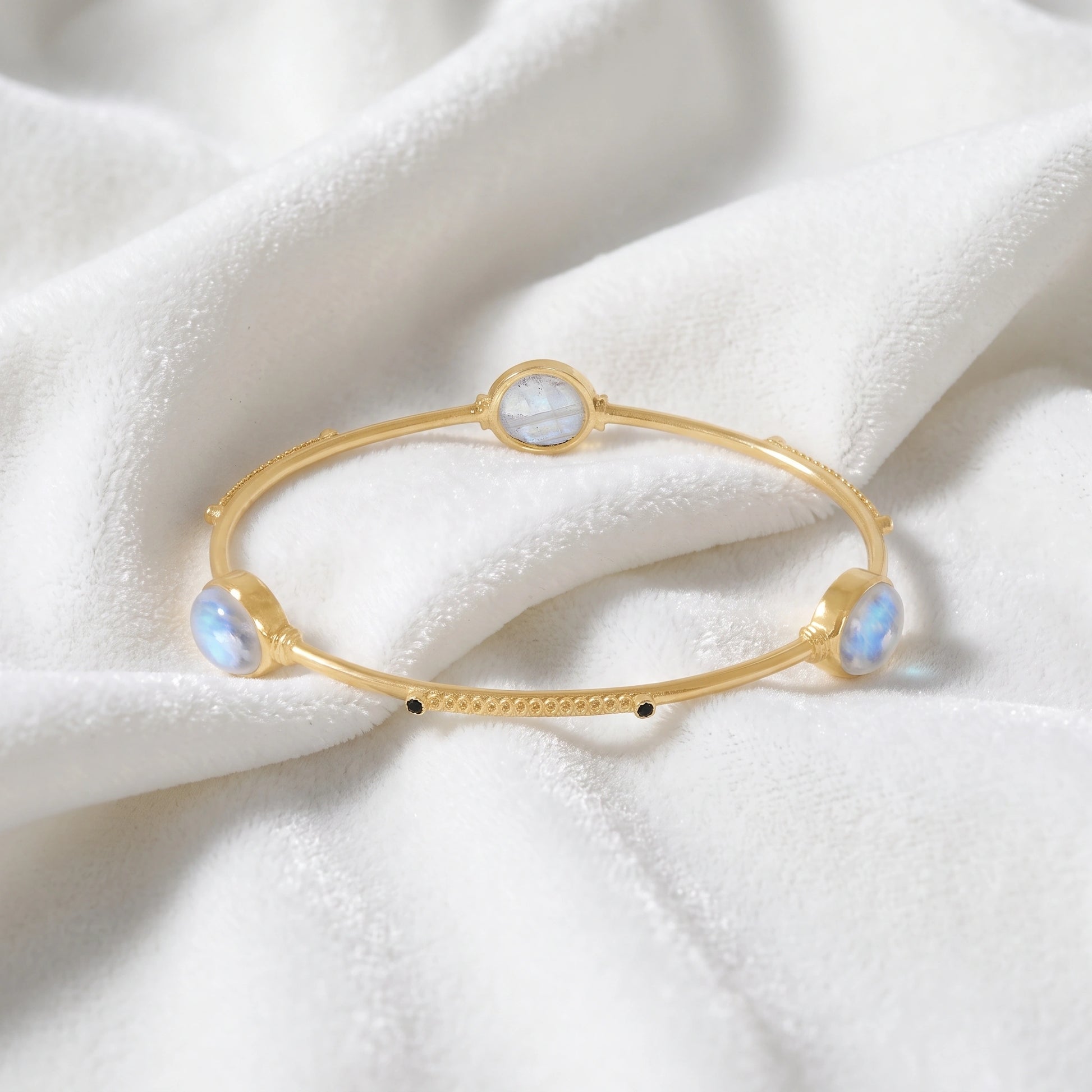 Bangle_bracelet_in_gold_plated_silver_with_oval_moonstones_and_black_onyx_stones_at_front_angle_view_on_soft_fabric_background
