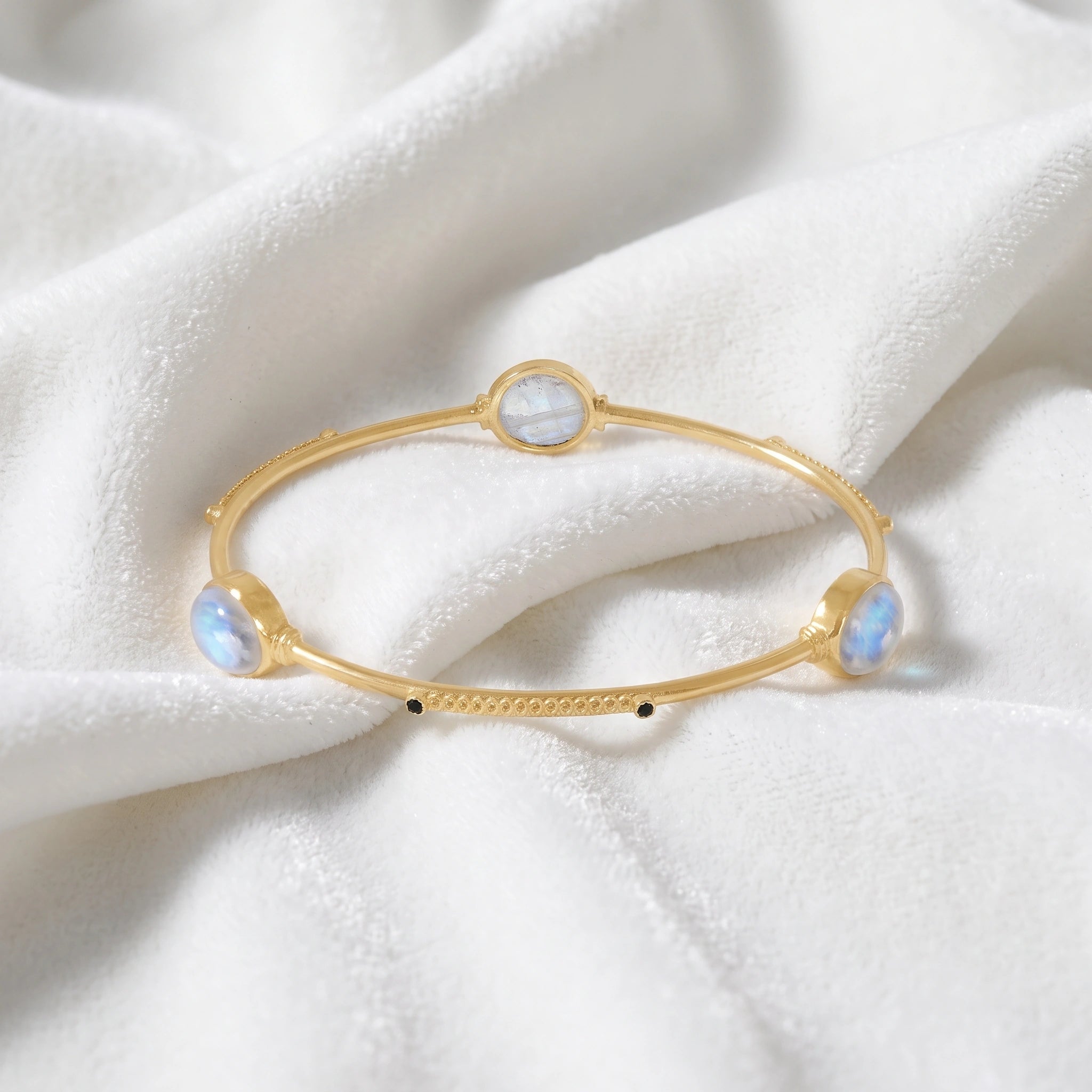 Bangle_bracelet_in_gold_plated_silver_with_oval_moonstones_and_black_onyx_stones_at_front_angle_view_on_soft_fabric_background