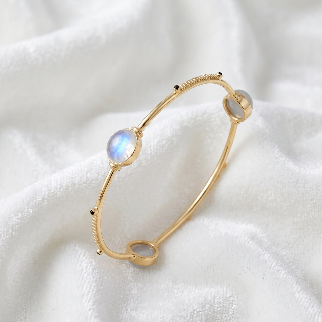 Bangle_bracelet_in_gold_plated_silver_with_oval_moonstones_and_black_onyx_stones_at_slight_angle_view_on_soft_fabric_background