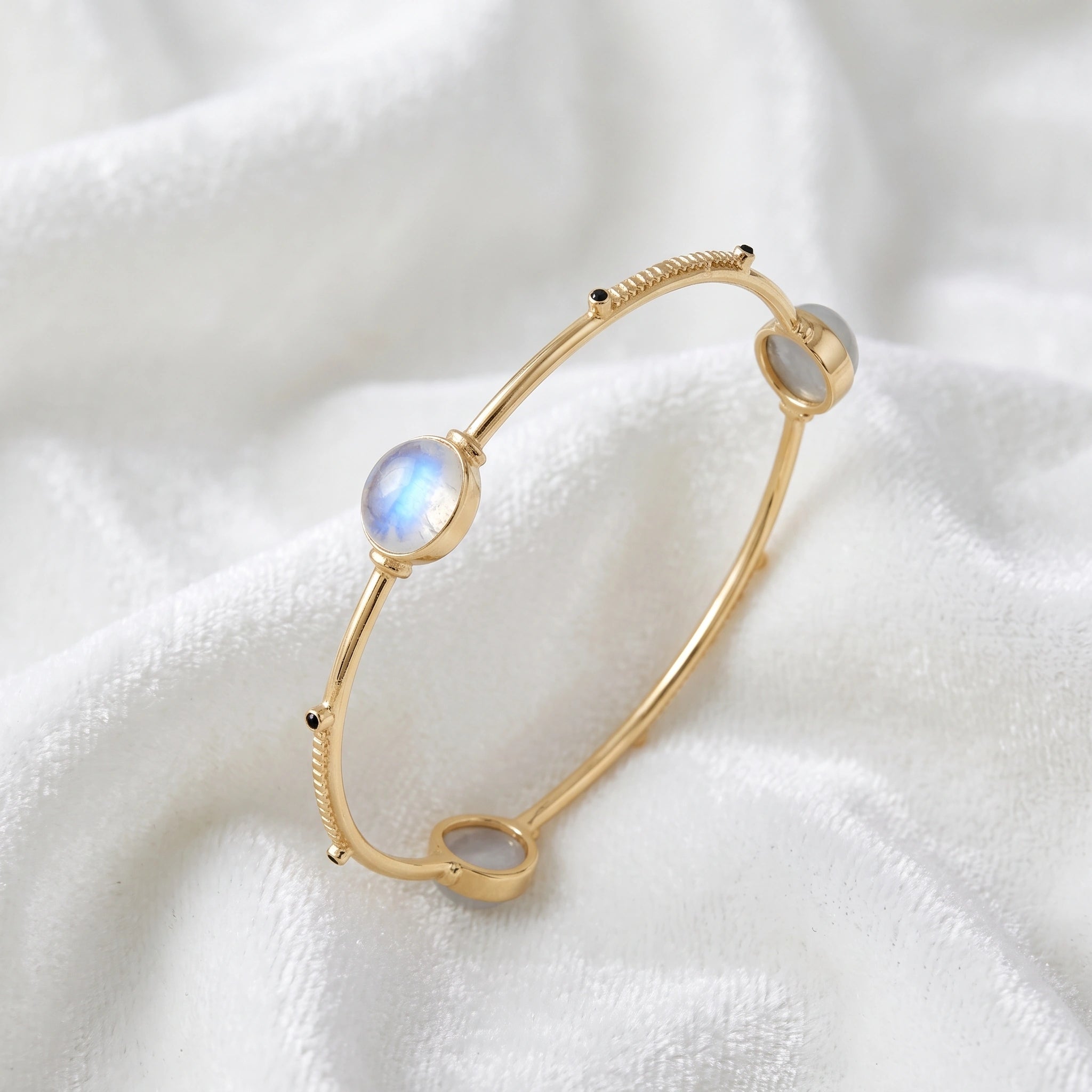 Bangle_bracelet_in_gold_plated_silver_with_oval_moonstones_and_black_onyx_stones_at_slight_angle_view_on_soft_fabric_background