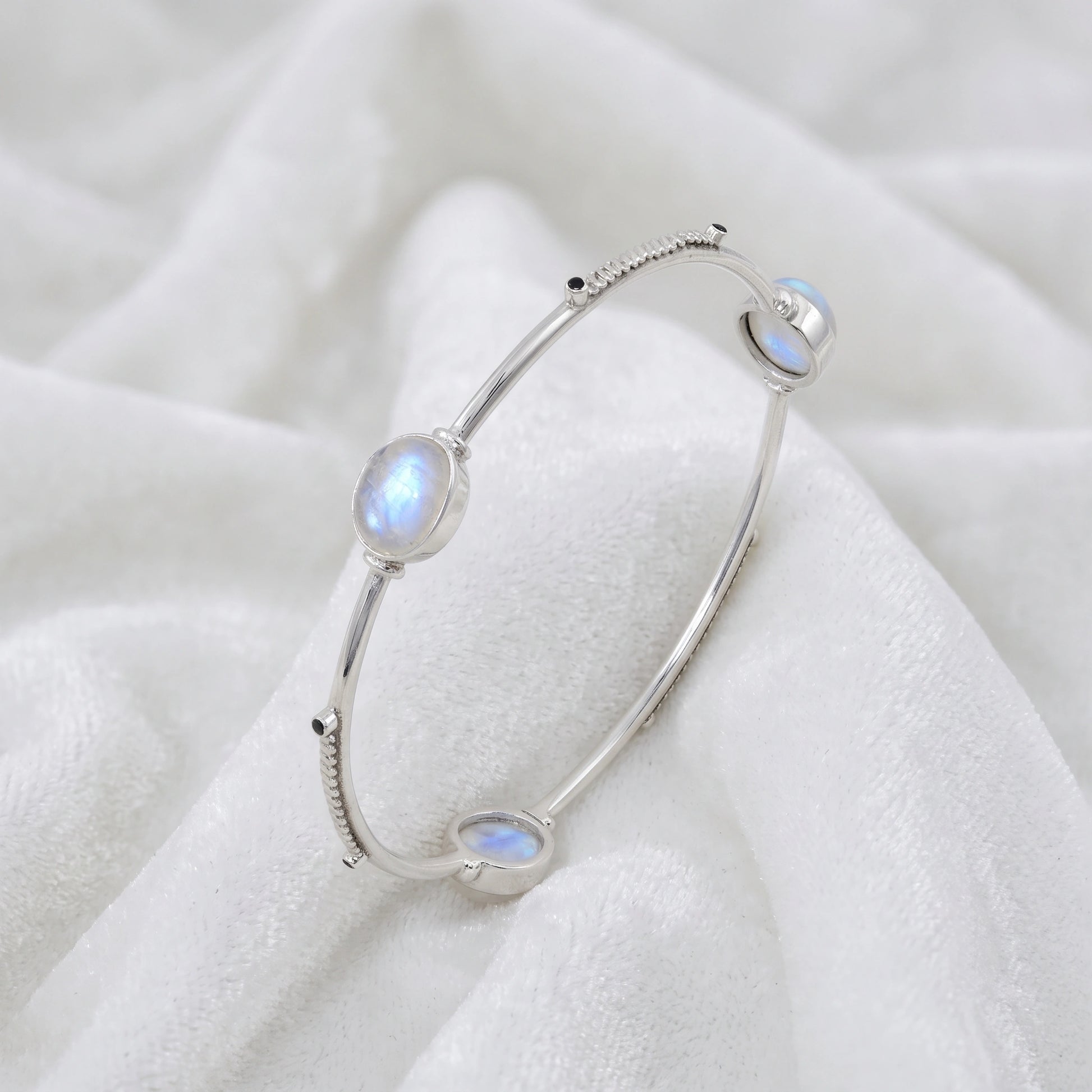 Bangle_bracelet_in_sterling_silver_with_oval_moonstones_and_black_onyx_stones_at_slight_angle_view_on_soft_fabric_background