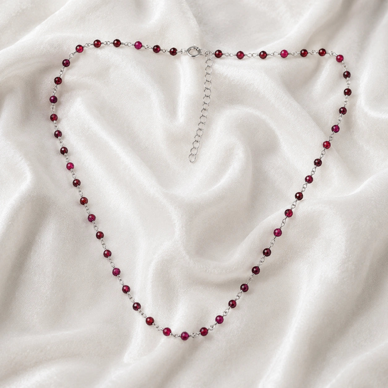 Beaded_necklace_with_sterling_silver_chain_and_deep_red_gemstone_beads_on_front_angle_view_on_soft_fabric_background