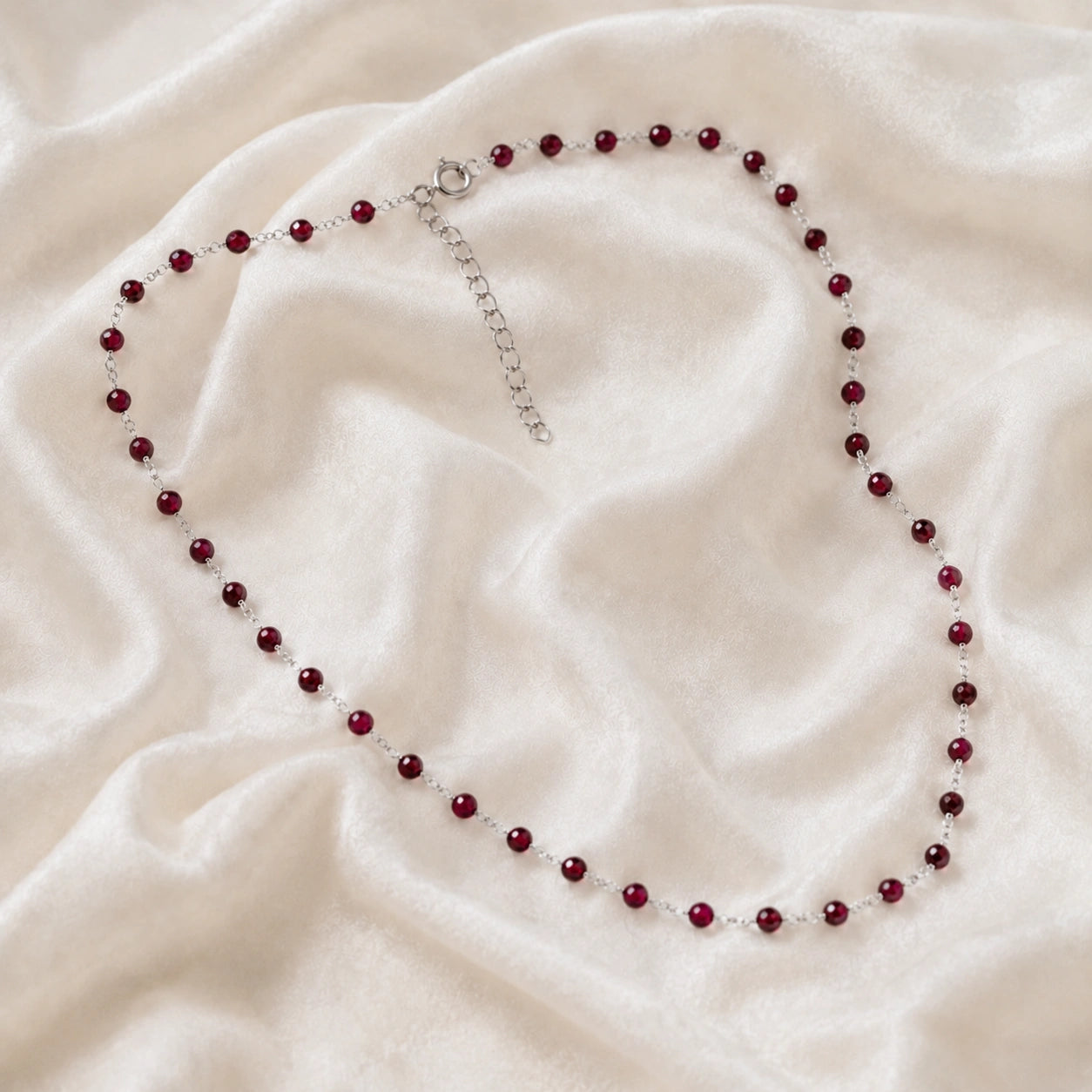 Beaded_necklace_with_sterling_silver_chain_and_deep_red_gemstone_beads_on_slightly_angled_view_on_soft_fabric_background