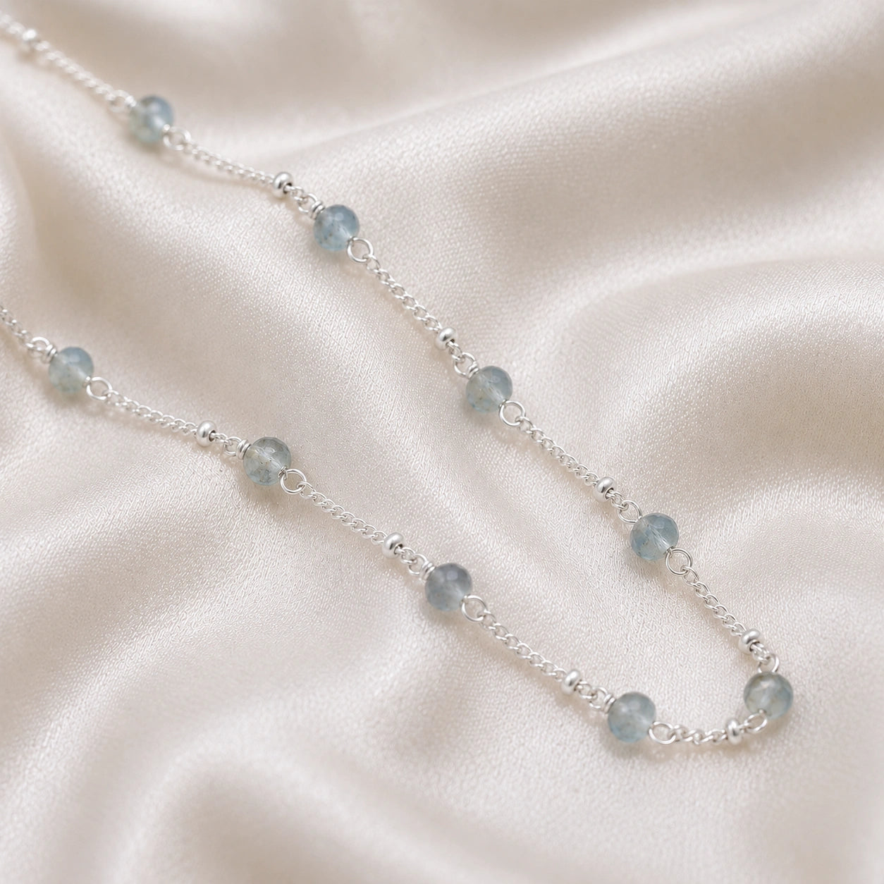 Beads_necklace_in_sterling_silver_at_slight_angle_with_aquamarine_stones_on_a_soft_fabric_background