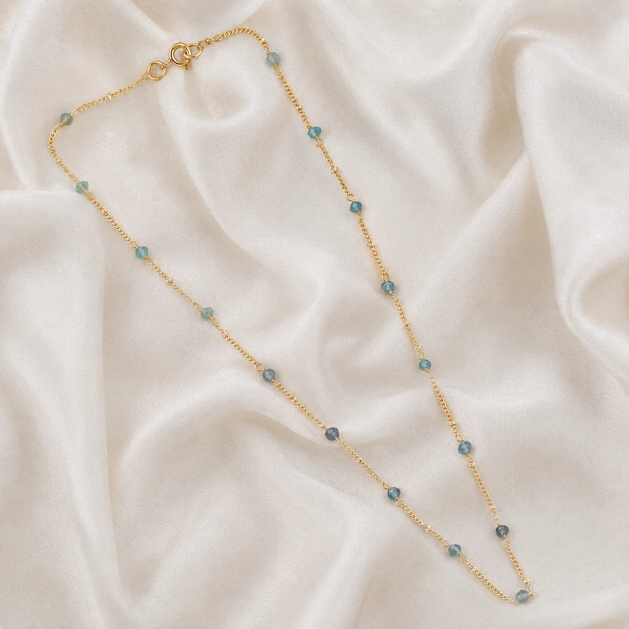 Beads_necklace_in_gold_plated_silver_at_front_angle_with_aquamarine_stones_on_a_soft_fabric_background