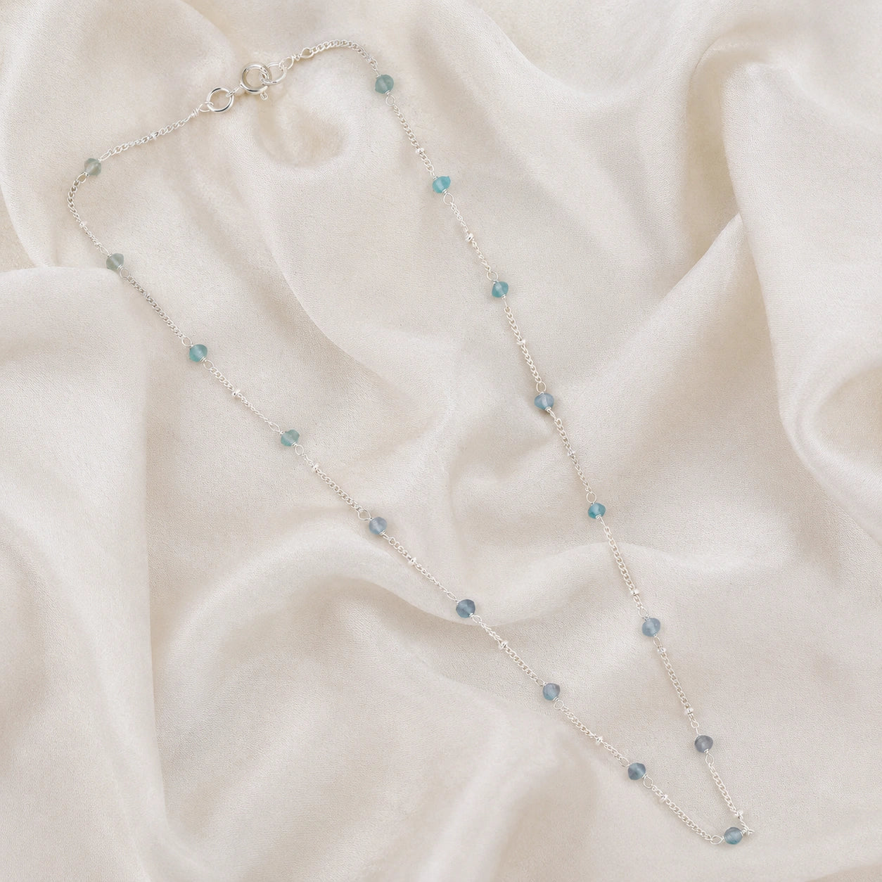 Beads_necklace_in_sterling_silver_at_front_angle_with_aquamarine_stones_on_a_soft_fabric_background