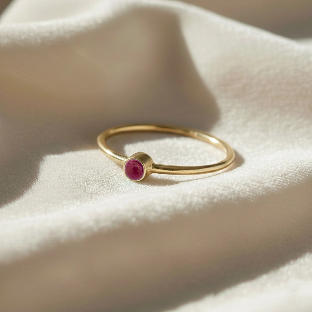 A gold plated birthstone ring featuring a ruby gemstone, styled as a delicate stacking ring on a soft white fabric background