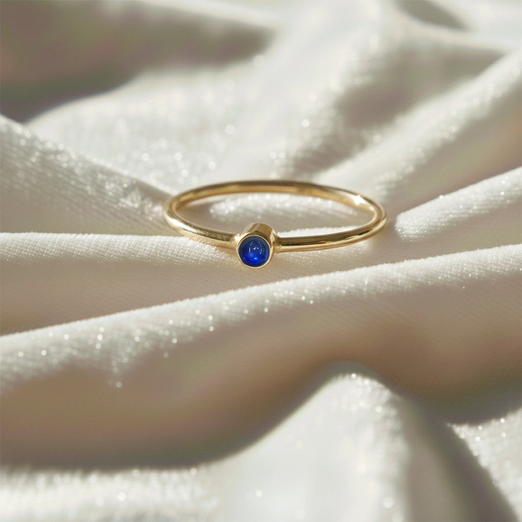 A gold plated birthstone ring featuring a sapphire gemstone, styled as a delicate stacking ring on a soft white fabric background