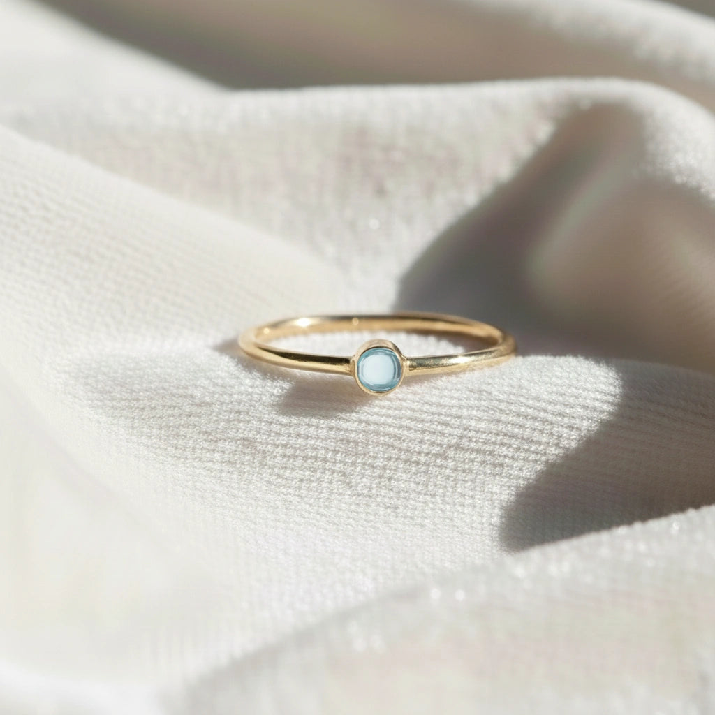 A gold plated birthstone ring featuring a blue topaz gemstone, styled as a delicate stacking ring on a soft white fabric background