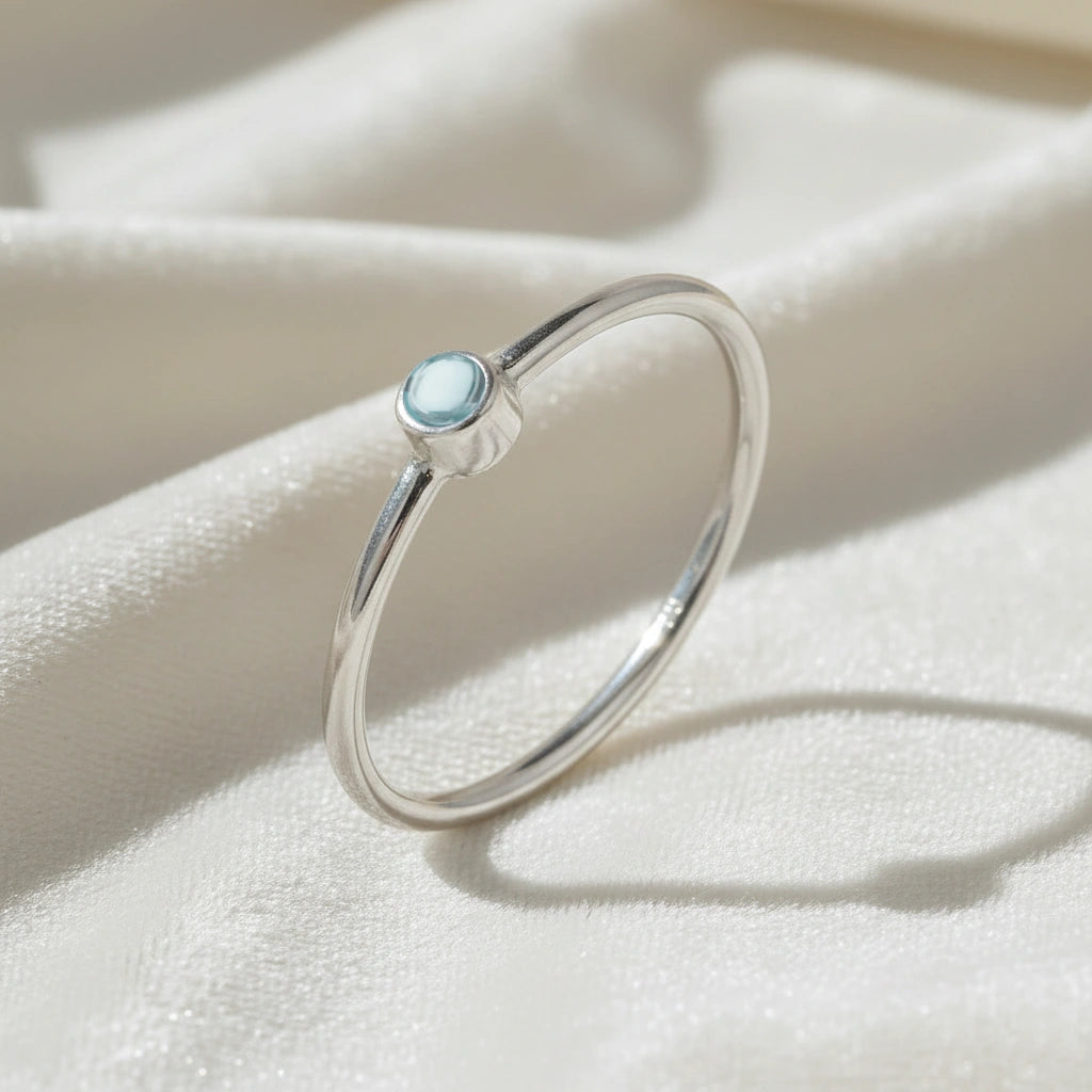 A silver birthstone ring featuring a blue topaz gemstone, styled as a delicate stacking ring on a soft white fabric background