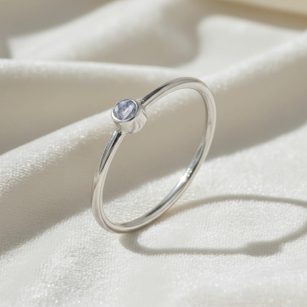 A silver birthstone ring featuring a clear quartz gemstone, styled as a delicate stacking ring on a soft white fabric background