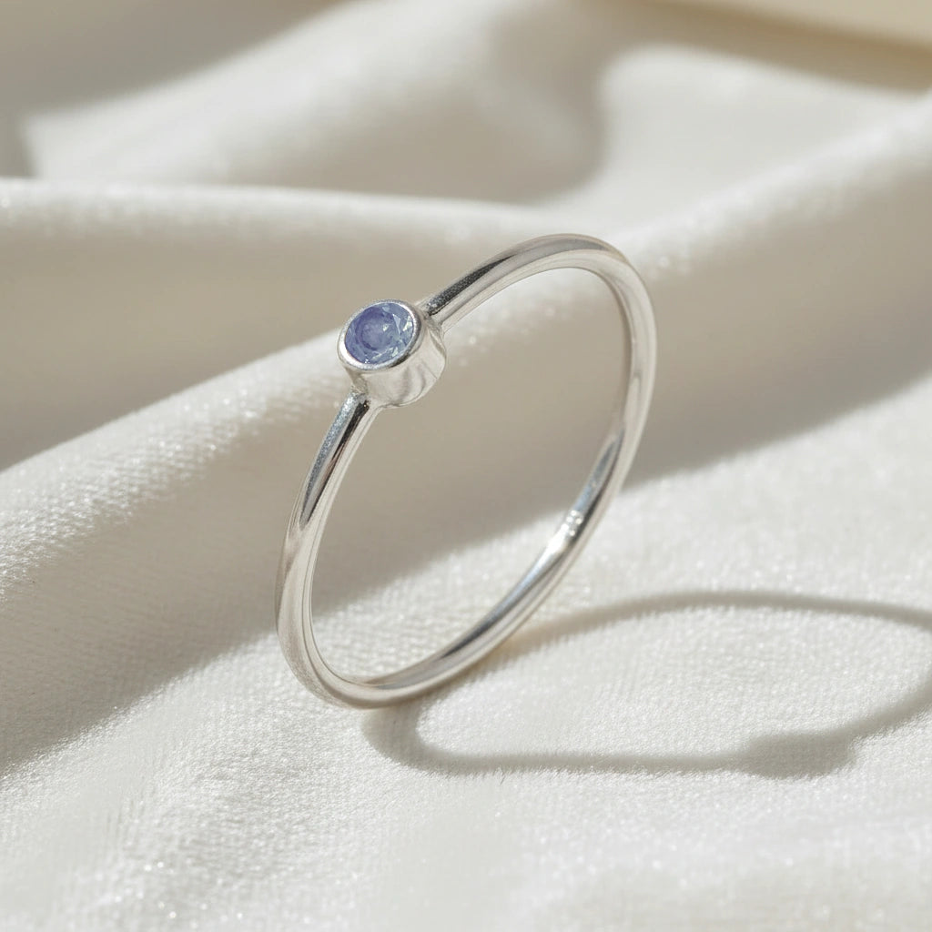 A silver birthstone ring featuring a tanzanite gemstone, styled as a delicate stacking ring on a soft white fabric background