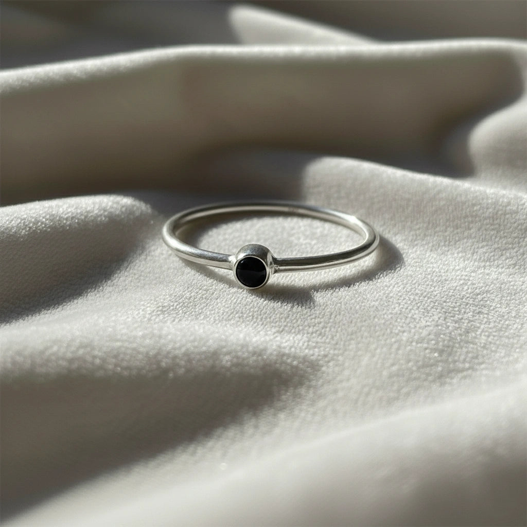 A silver birthstone ring featuring a black onyx gemstone, styled as a delicate stacking ring on a soft white fabric background