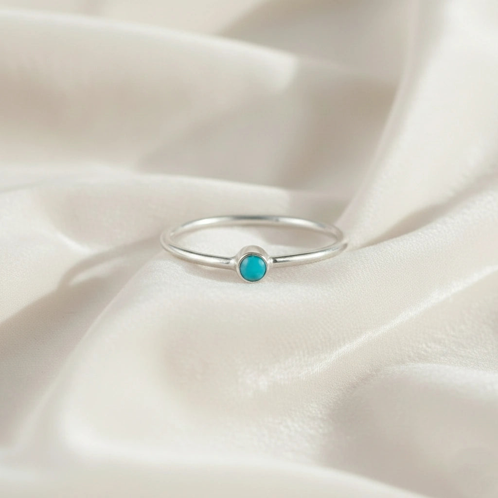A silver birthstone ring featuring a turquoise gemstone, styled as a delicate stacking ring on a soft white fabric background