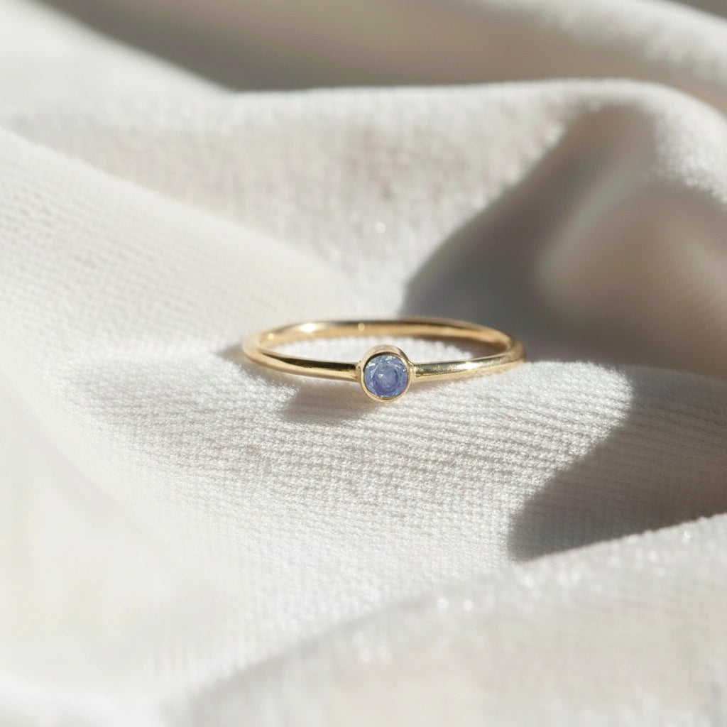 A gold plated birthstone ring featuring a tanzanite gemstone, styled as a delicate stacking ring on a soft white fabric background