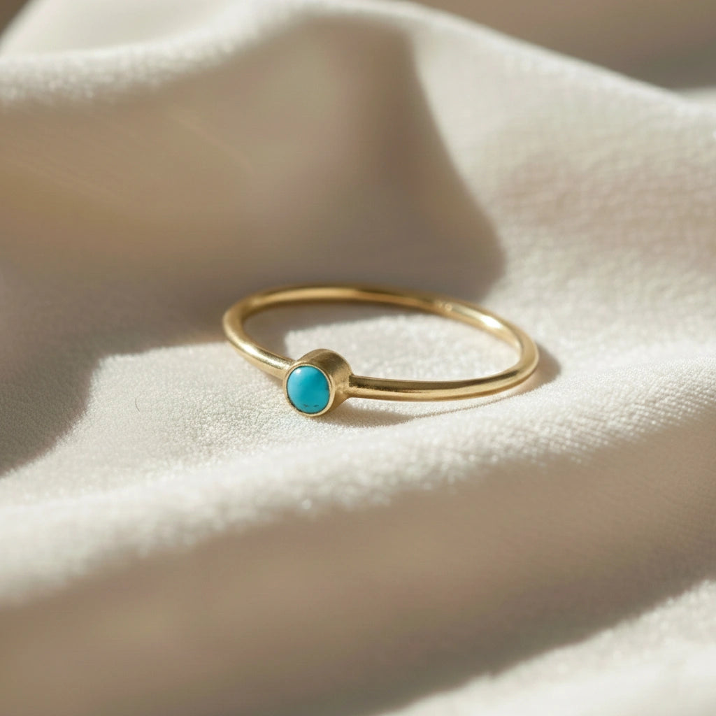 A gold plated birthstone ring featuring a turquoise gemstone, styled as a delicate stacking ring on a soft white fabric background