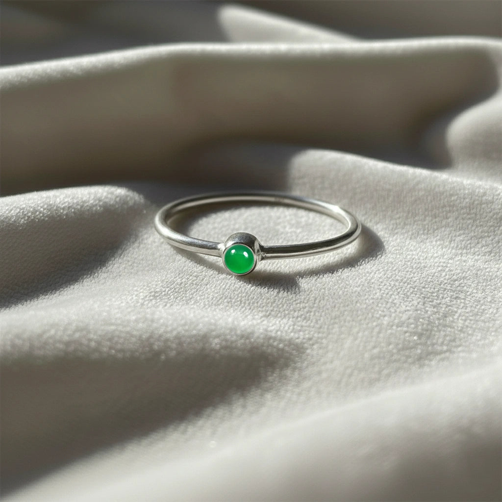 A silver birthstone ring featuring a green onyx gemstone, styled as a delicate stacking ring on a soft white fabric background