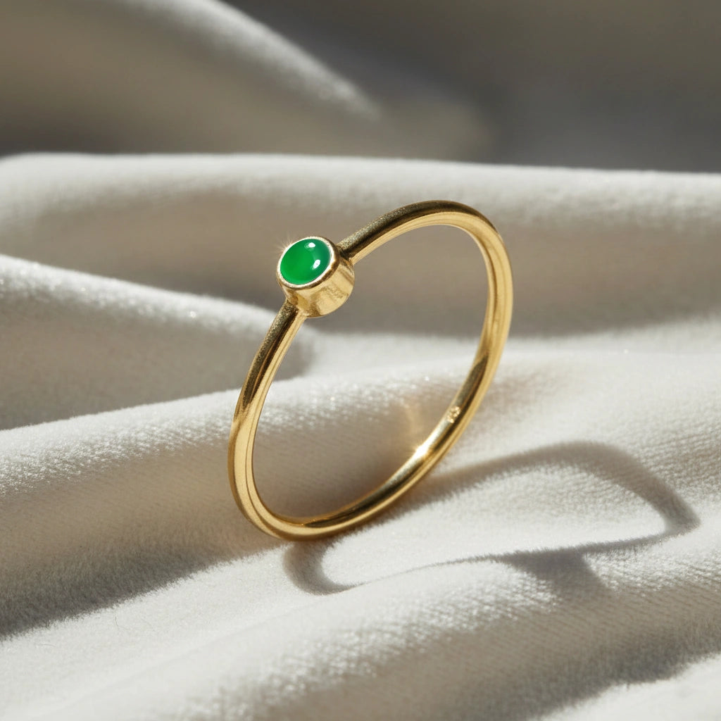 A gold plated birthstone ring featuring a green onyx gemstone, styled as a delicate stacking ring on a soft white fabric background