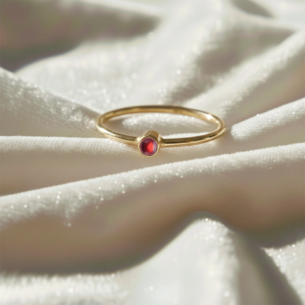 A gold plated birthstone ring featuring a garnet gemstone, styled as a delicate stacking ring on a soft white fabric background
