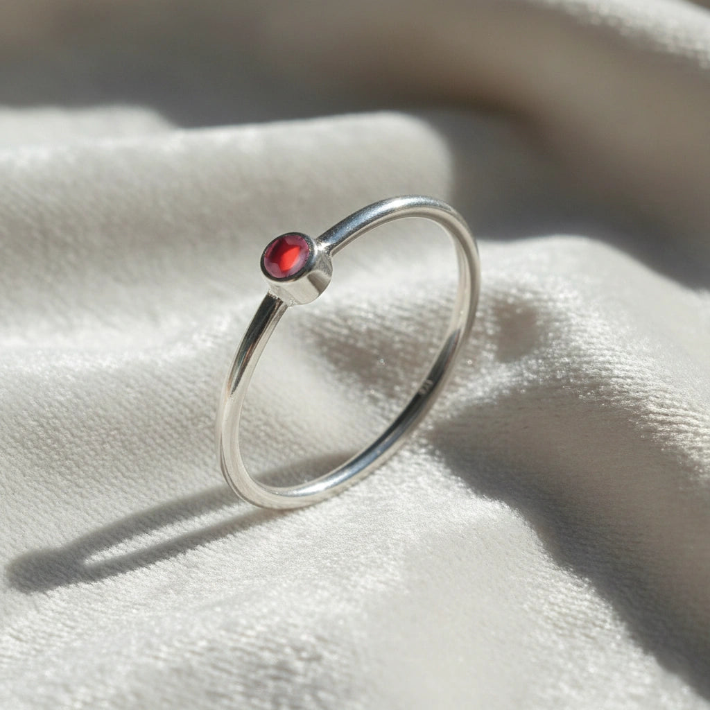 A silver birthstone ring featuring a garnet gemstone, styled as a delicate stacking ring on a soft white fabric background