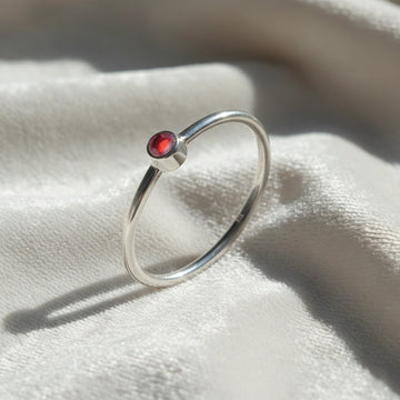 A silver birthstone ring featuring a garnet gemstone, styled as a delicate stacking ring on a soft white fabric background