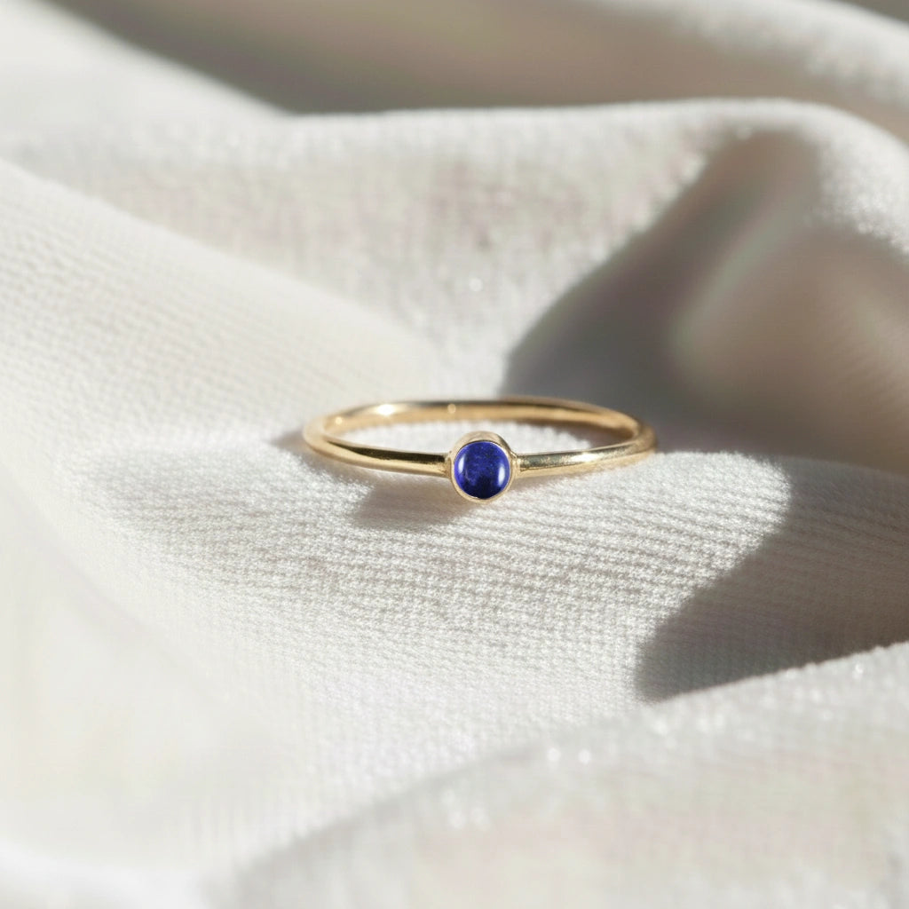 A gold plated birthstone ring featuring a lapis lazuli gemstone, styled as a delicate stacking ring on a soft white fabric background