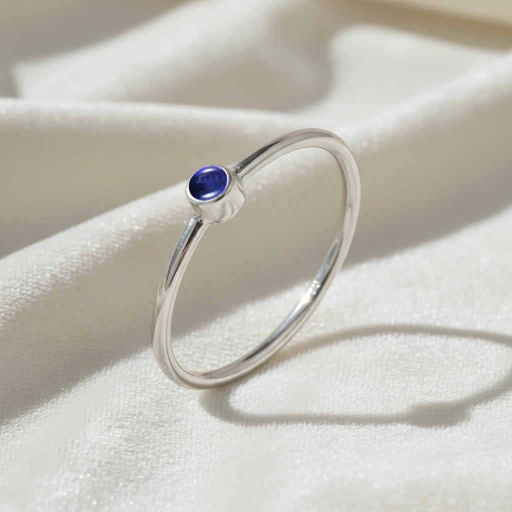 A silver birthstone ring featuring a lapis lazuli gemstone, styled as a delicate stacking ring on a soft white fabric background