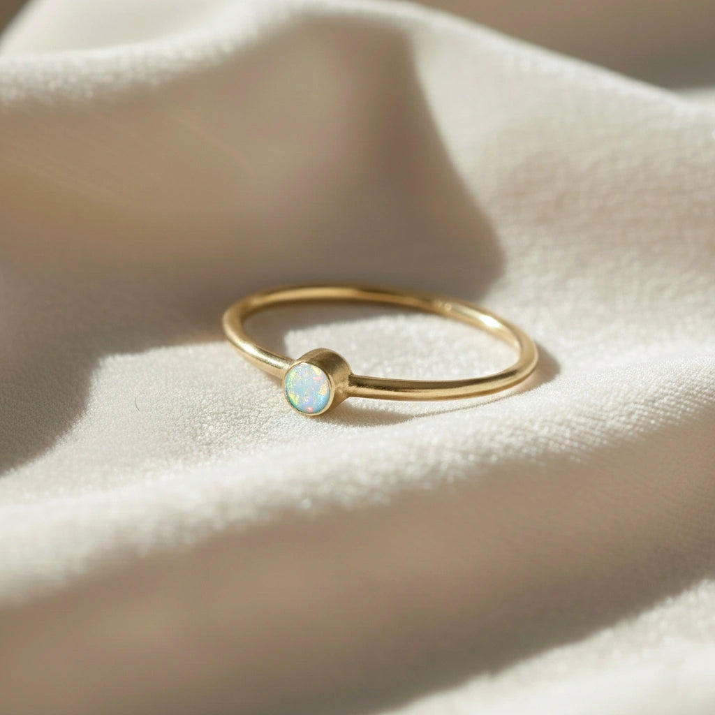 A gold plated birthstone ring featuring an opal gemstone, styled as a delicate stacking ring on a soft white fabric background