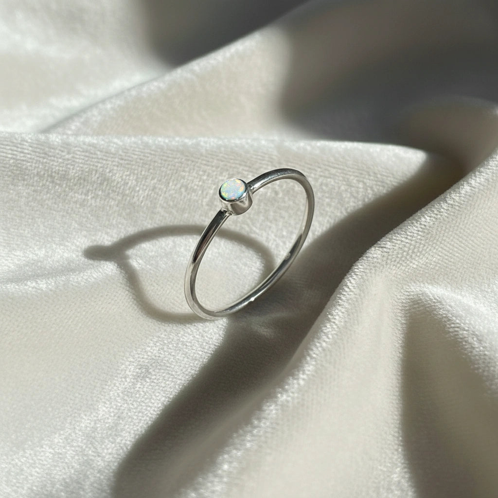 A silver birthstone ring featuring an opal gemstone, styled as a delicate stacking ring on a soft white fabric background