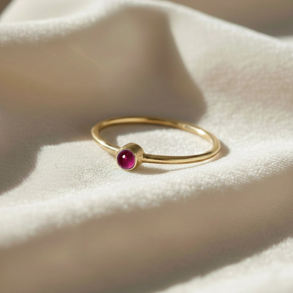 A gold plated birthstone ring featuring a pink tourmaline gemstone, styled as a delicate stacking ring on a soft white fabric background