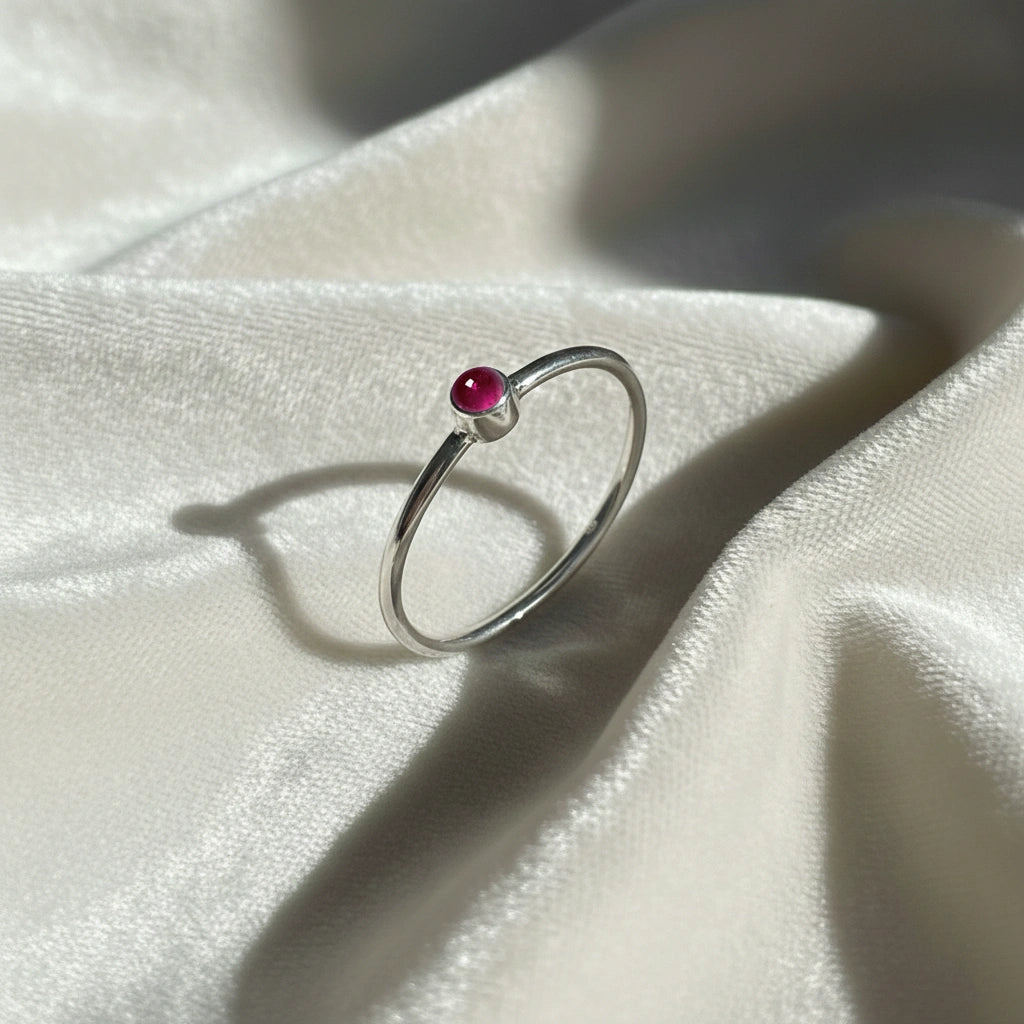 A silver birthstone ring featuring a pink tourmaline gemstone, styled as a delicate stacking ring on a soft white fabric background