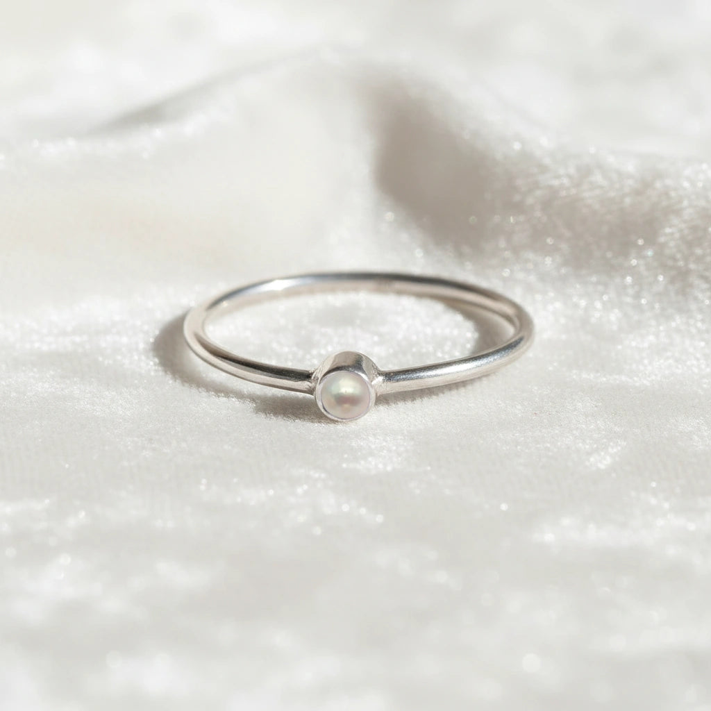A silver birthstone ring featuring a pearl  gemstone, styled as a delicate stacking ring on a soft white fabric background