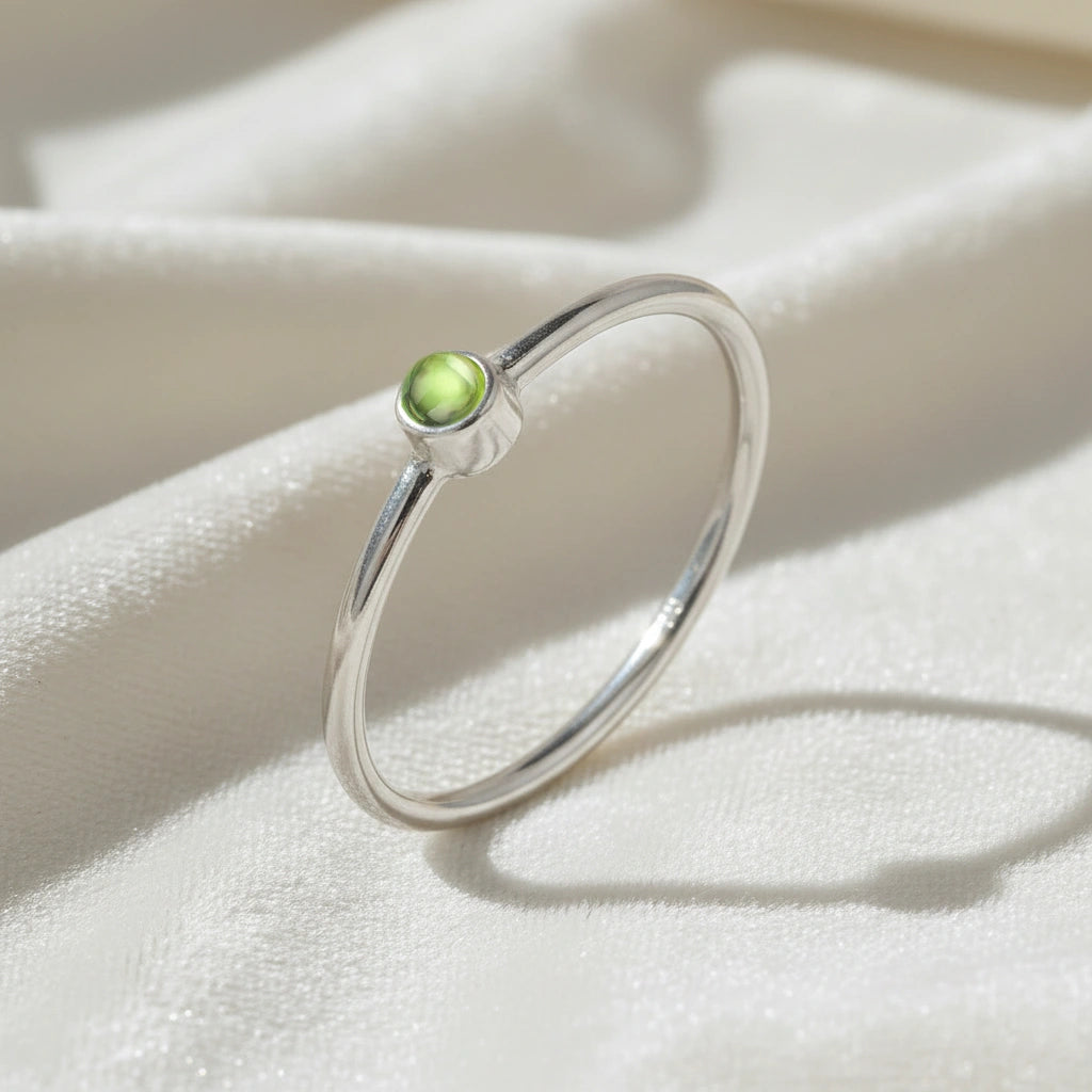 A silver birthstone ring featuring a peridot gemstone, styled as a delicate stacking ring on a soft white fabric background