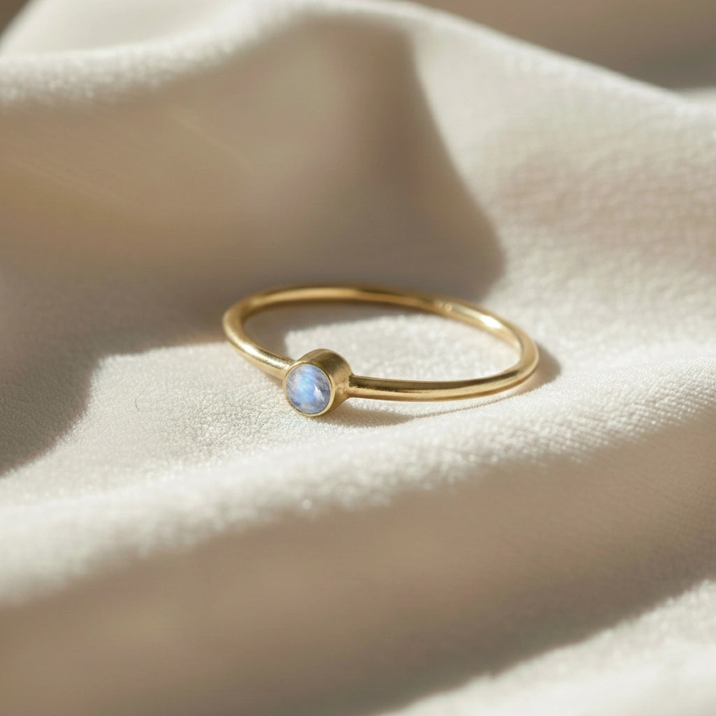 A gold plated birthstone ring featuring a moonstone gemstone, styled as a delicate stacking ring on a soft white fabric background