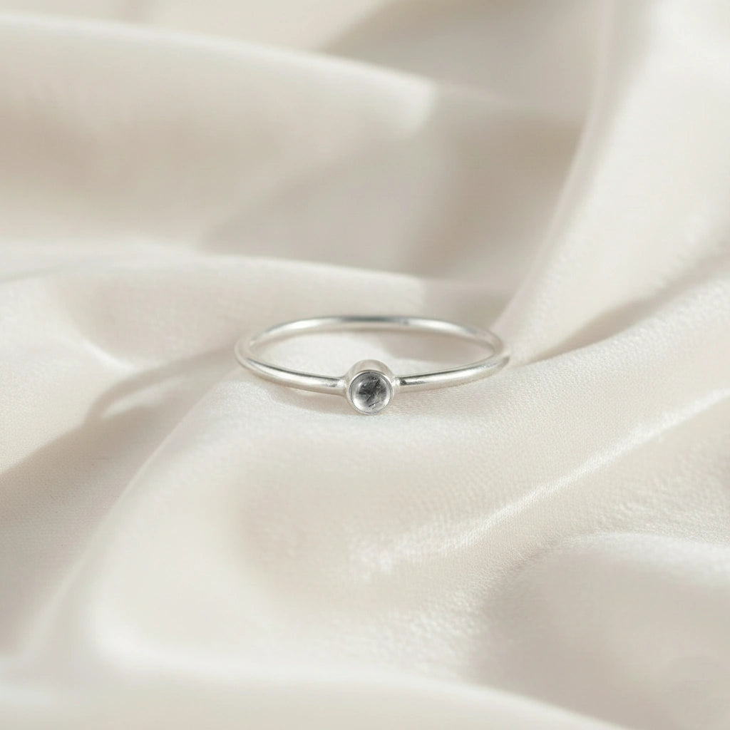 A silver birthstone ring featuring a white topaz gemstone, styled as a delicate stacking ring on a soft white fabric background