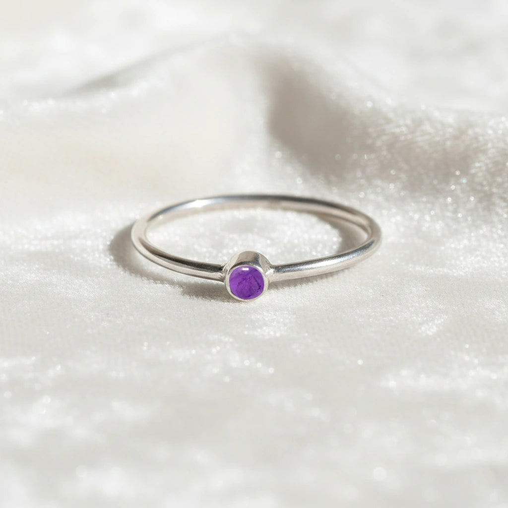 A silver birthstone ring featuring a amethyst gemstone, styled as a delicate stacking ring on a soft white fabric background