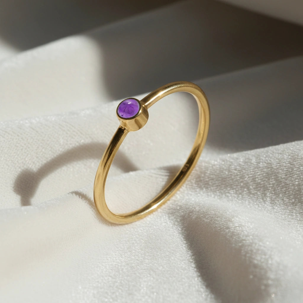 A gold plated birthstone ring featuring a amethyst gemstone, styled as a delicate stacking ring on a soft white fabric background