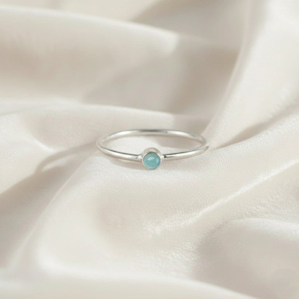A silver birthstone ring featuring a aquamarine gemstone, styled as a delicate stacking ring on a soft white fabric background