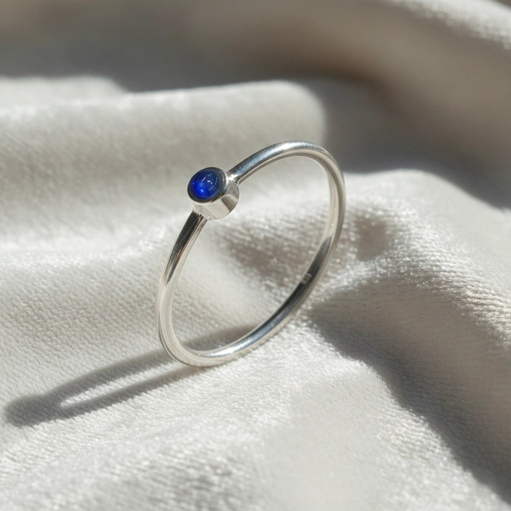 A silver birthstone ring featuring a sapphire gemstone, styled as a delicate stacking ring on a soft white fabric background